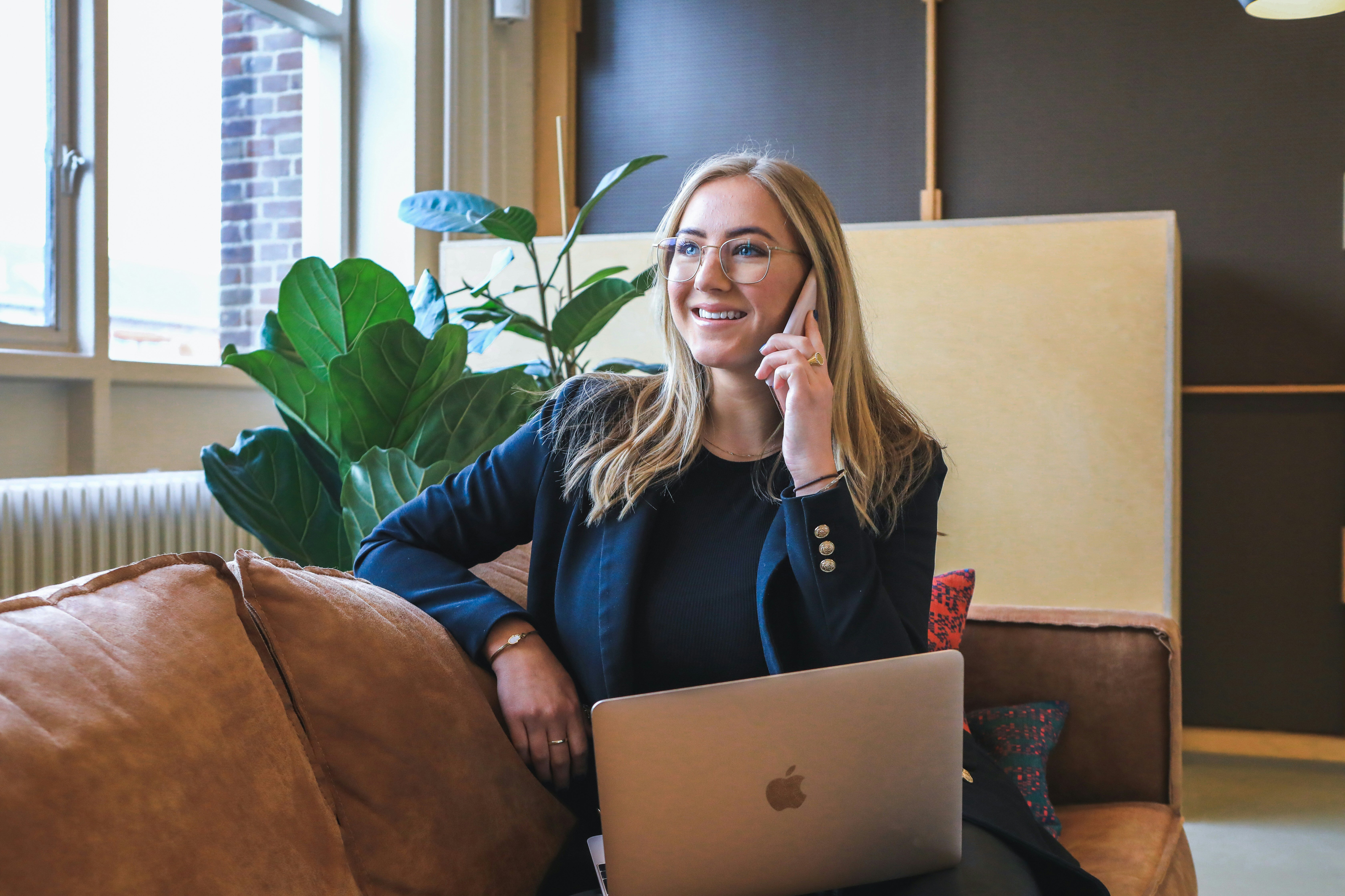 woman in professional attire sitting on a couch with her laptop open and talking on the phone