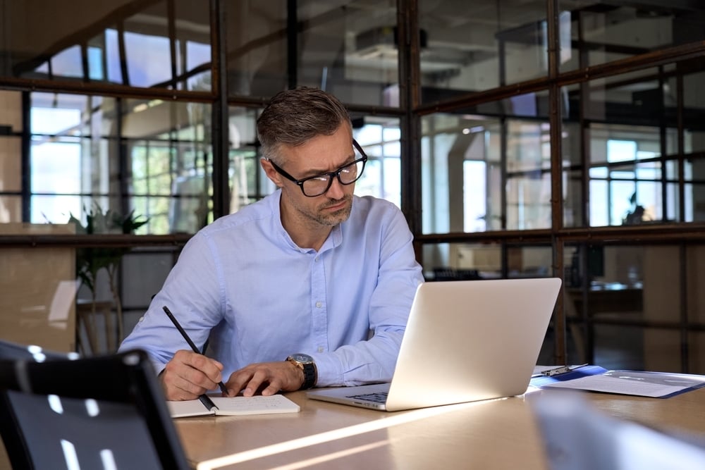 man working at desk with laptop open writing in a notebook 